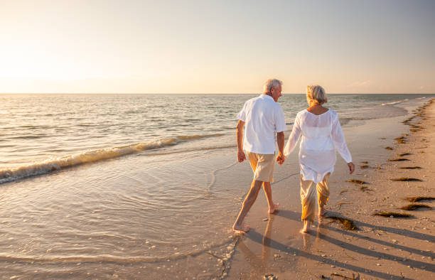 feliz pareja de ancianos jubilados caminando tomados de la mano en la playa al atardecer